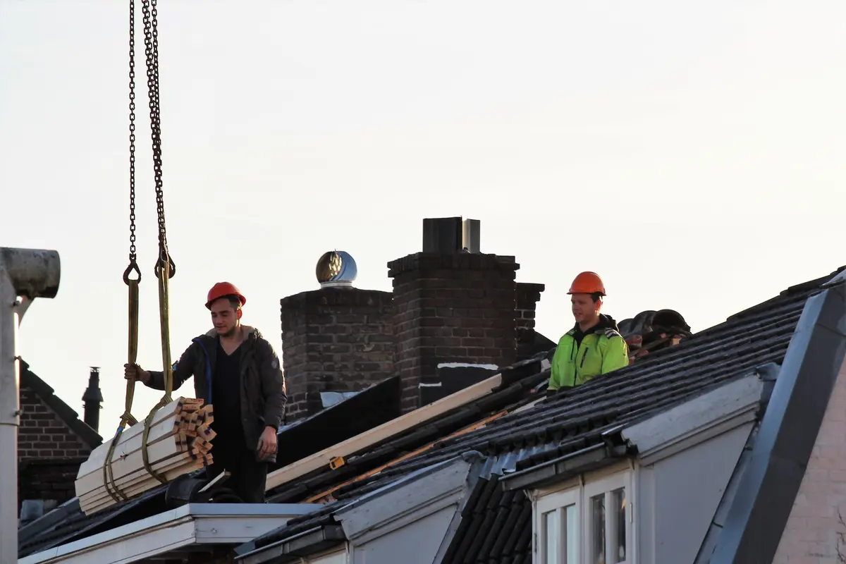 Twee dakdekkers met helm op het dak van Nederlandse rijtjeshuizen, met balken aan een kraan